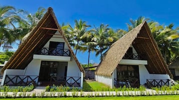 Deux bungalows aux toits de chaume, dotés de balcons, entourés de palmiers et d’une pelouse bien entretenue.