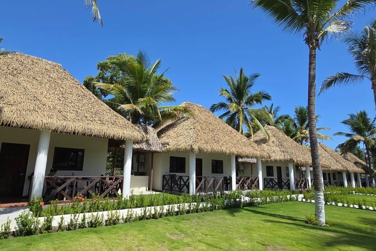 A row of thatched-roof cottages with white walls and wooden railings, surrounded by well-maintained grass and palm trees.