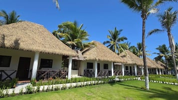 Una hilera de cabañas con techo de paja, paredes blancas y barandillas de madera, rodeadas de césped cuidado y palmeras.