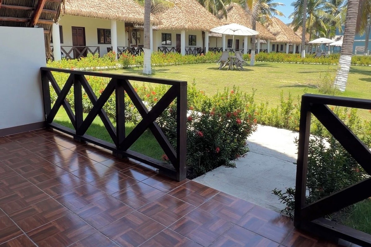 A covered outdoor area with a wooden floor, a thatched roof, and a view of a resort with palm trees and a clear sky.