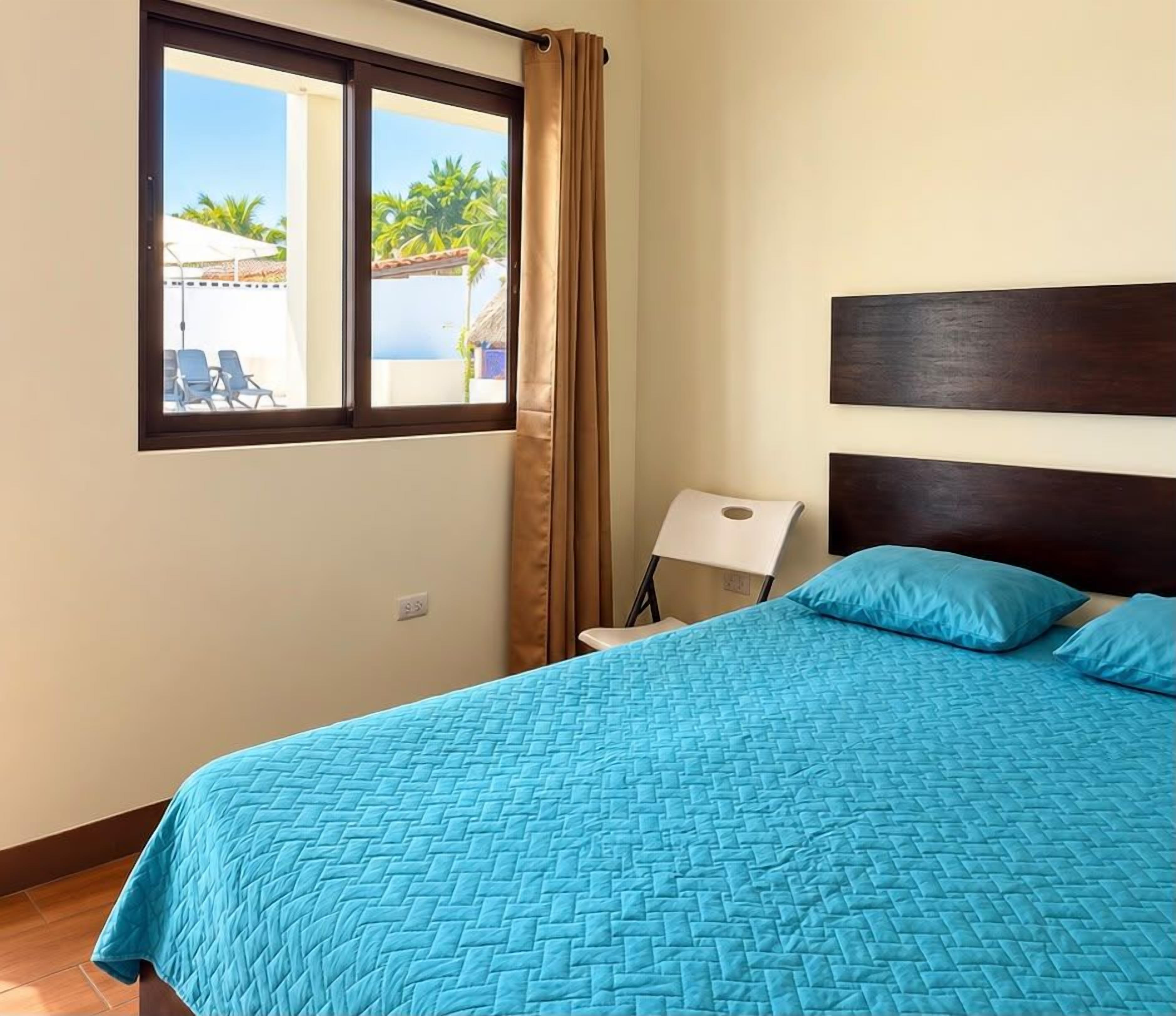 A room with a bed, a window with curtains, a chair, and a view of a pool and palm trees.