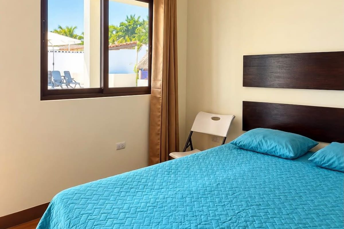 A room with a bed, a window with curtains, a chair, and a view of a pool and palm trees.