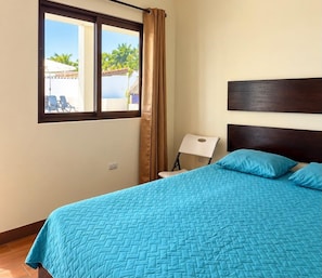 A room with a bed, a window with curtains, a chair, and a view of a pool and palm trees. - Hotel El Marinero (Chiquimulilla)