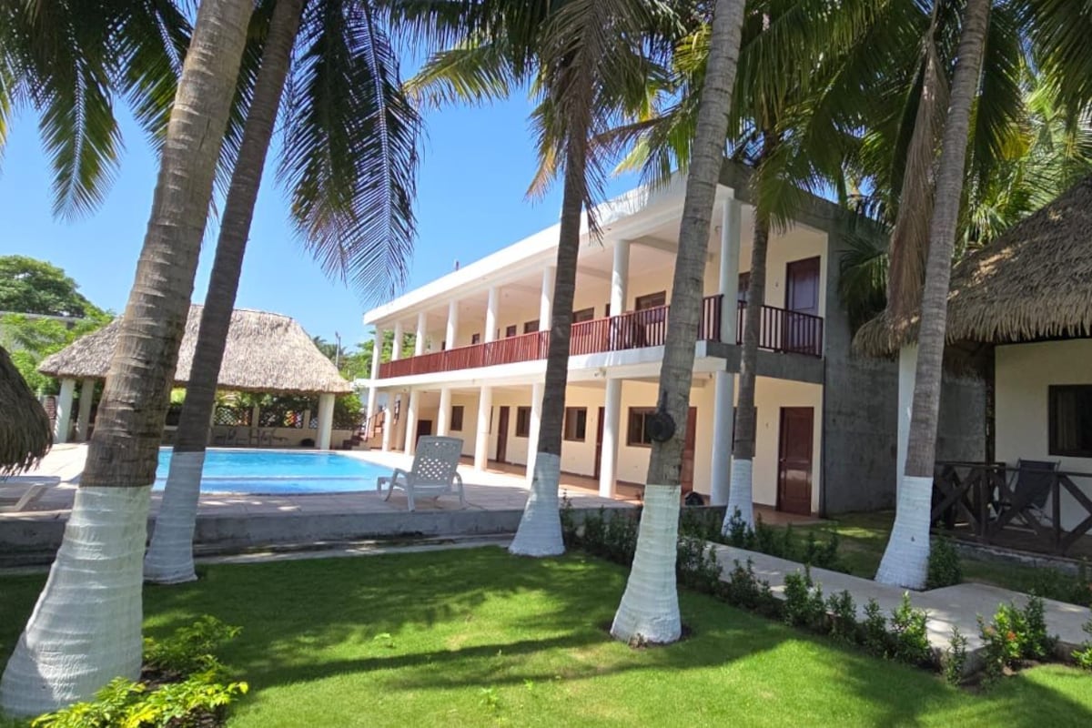 A resort with a pool, palm trees, and a thatched-roof building.