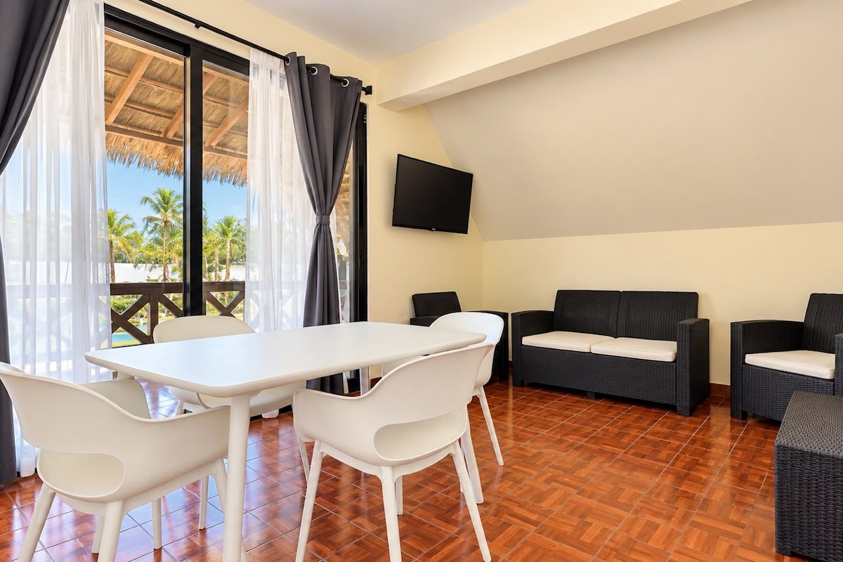 A room with a dining table and chairs, a sofa, and a TV on the wall.