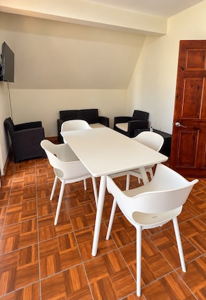 A dining area with a white table and chairs, a wooden herringbone floor, a television mounted on the wall, and a wooden door.