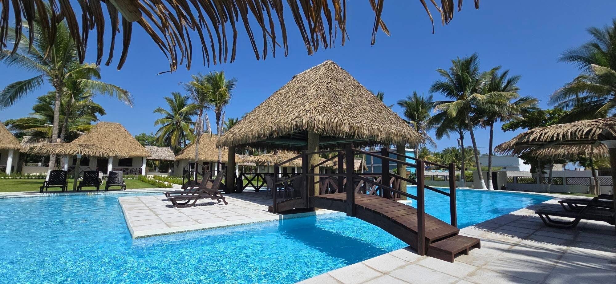 Un área de piscina con estructuras de techo de paja, sillas de descanso y palmeras.