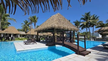 Un área de piscina con estructuras de techo de paja, sillas de descanso y palmeras.
