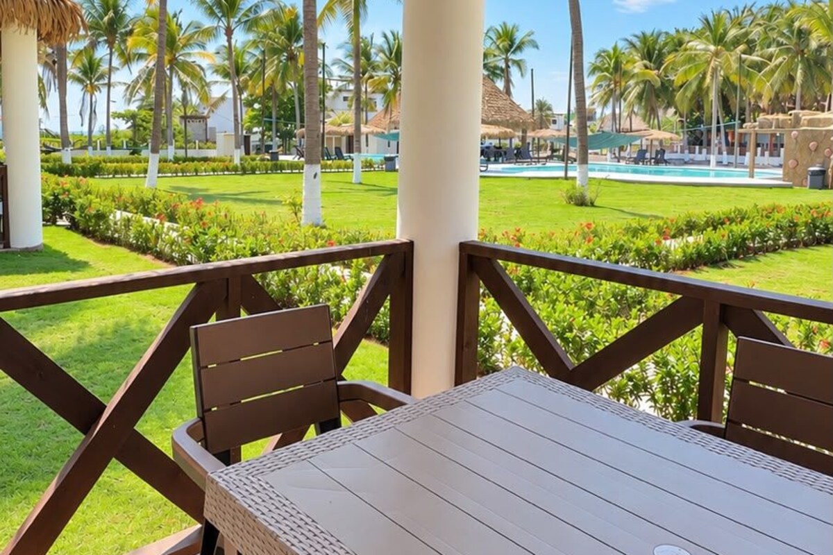 A covered outdoor seating area with a table and chairs, overlooking a pool and palm trees.