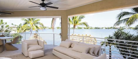 A sunlit room with a ceiling fan, a sofa, a table, and a view of a body of water with palm trees.