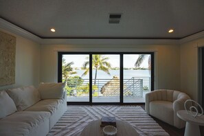 Quarto com sofá branco, poltrona branca, mesa branca com um livro e vista para um corpo d'água com palmeiras.