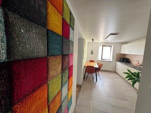 Eine moderne Küche mit weißen Schränken, ein Essbereich mit einem Holztisch und orangefarbenen Stühlen sowie eine farbenfrohe Wand.