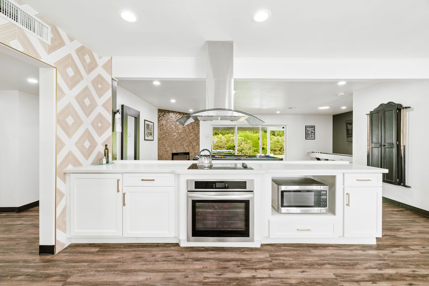 A modern kitchen with white cabinetry, a central island, a built-in oven, and a view of greenery outside.