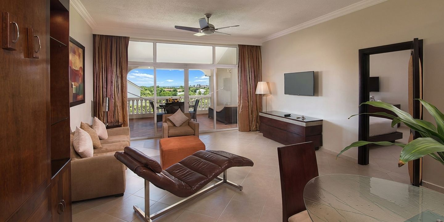 Ein Hotelzimmer mit Sofa, Liegekorb, Glastisch, Fernseher und einem Balkon mit Blick.