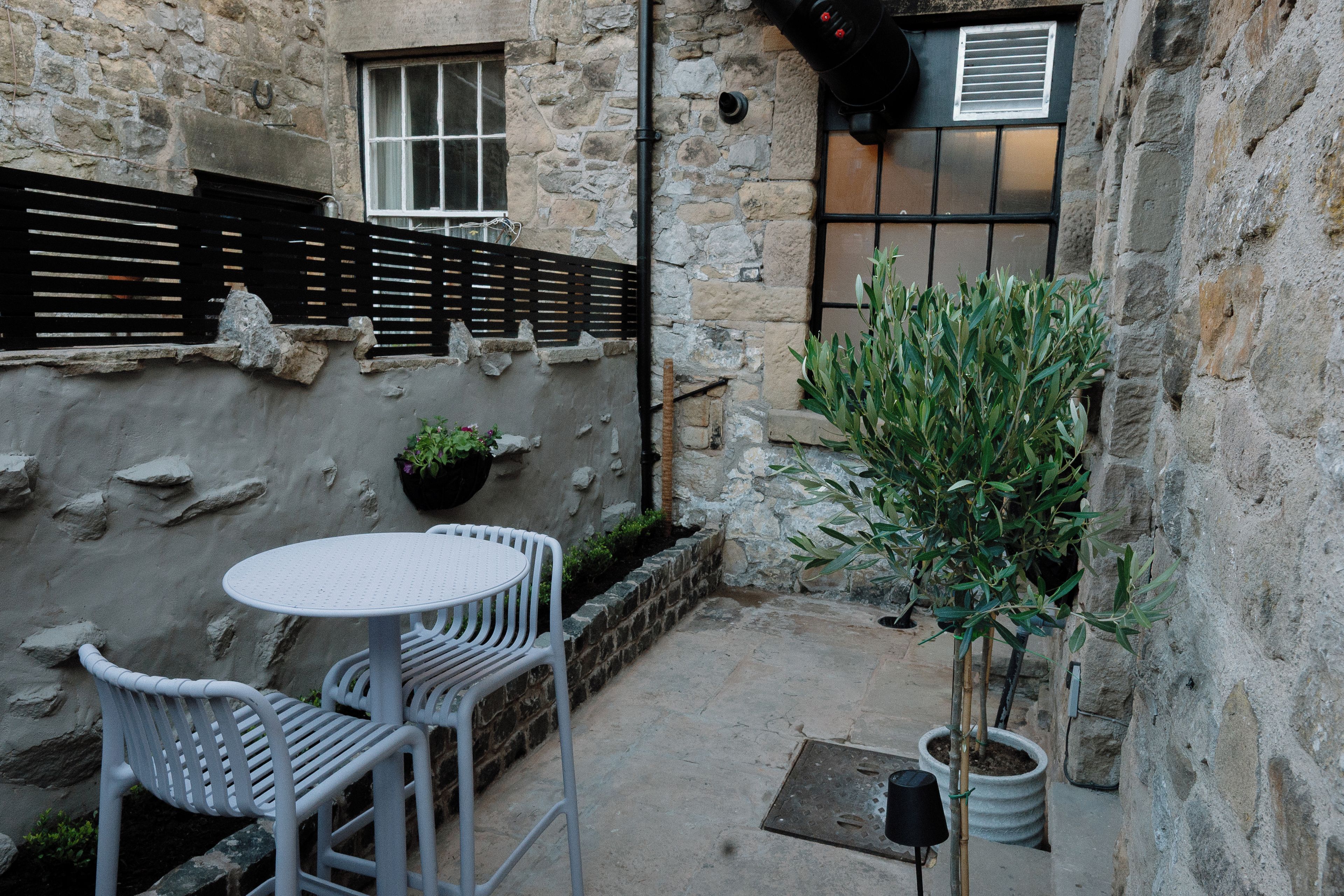 Une cour pavée de pierres, avec une table ronde et deux chaises, un pot de fleurs et un petit arbre.