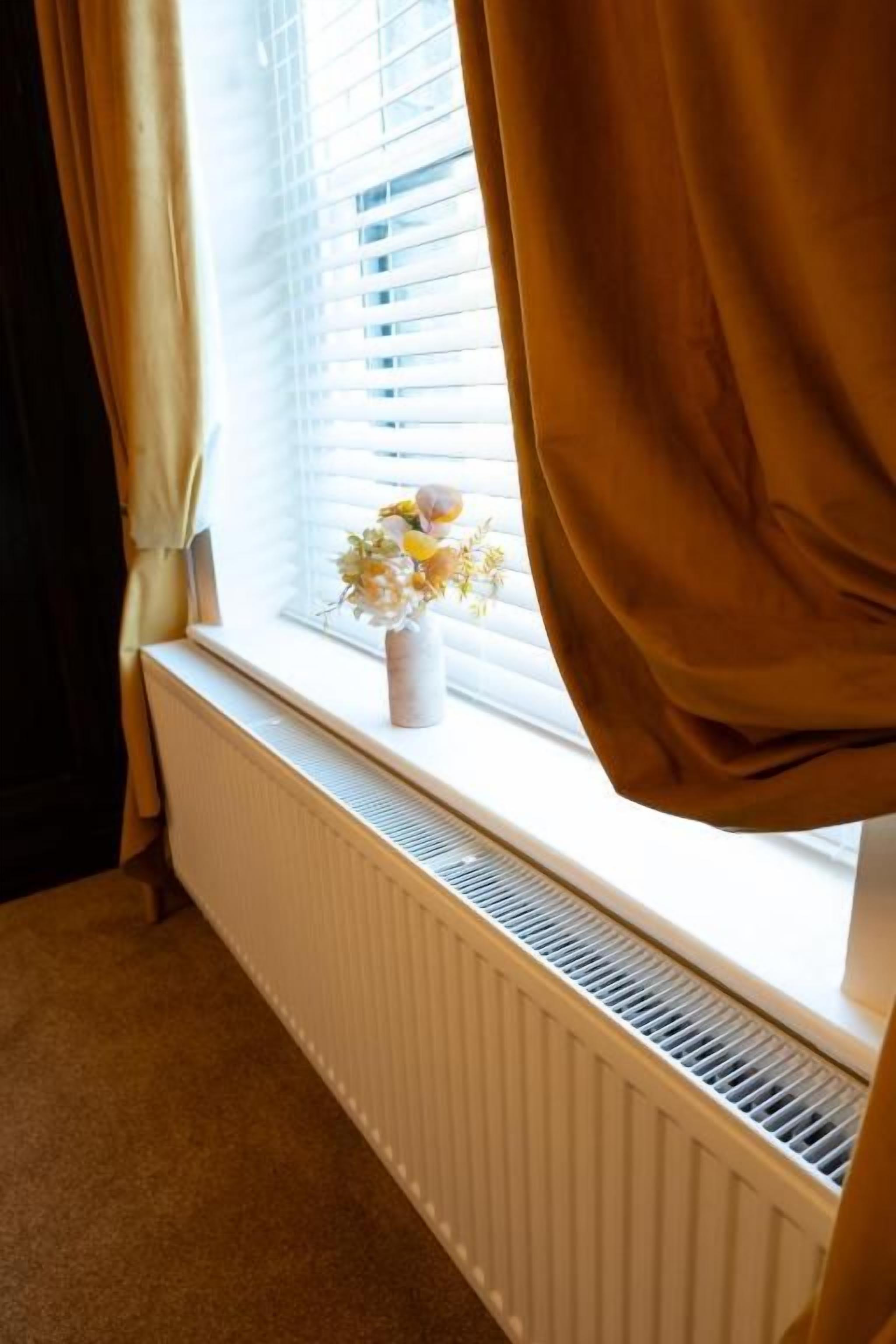 A room with a radiator, a window with blinds, and a vase with flowers on the sill.