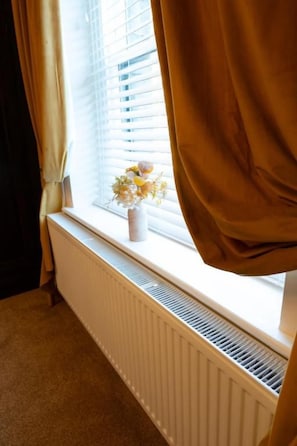 A room with a radiator, a window with blinds, and a vase with flowers on the sill. - UpBuxton (Buxton)