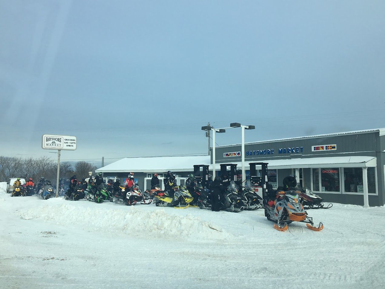 A snow-covered outdoor area with a building labeled "Bay Shore Market" and several snowmobiles parked in front.