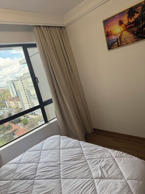 A bedroom with a bed, a window with curtains, a painting of a sunset, and a view of buildings outside. - Gorgeous 1-bedroom apartment in Nairobi kilimani perfect for your stay (Nairobi)