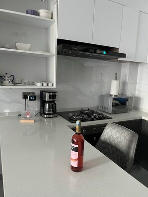 Una cucina moderna con mobili bianchi, piano cottura in acciaio inossidabile, una macchina da caffè e una bottiglia di vino rosso sul bancone.