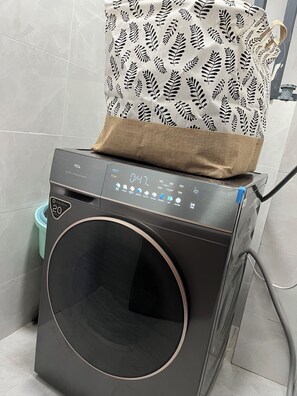 Una lavatrice a carica frontale con pannello di controllo, un cestino della spazzatura blu e una borsa fantasia sopra.