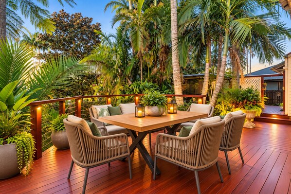 An outdoor patio with a wooden table and wicker chairs, surrounded by lush tropical plants and palm trees.