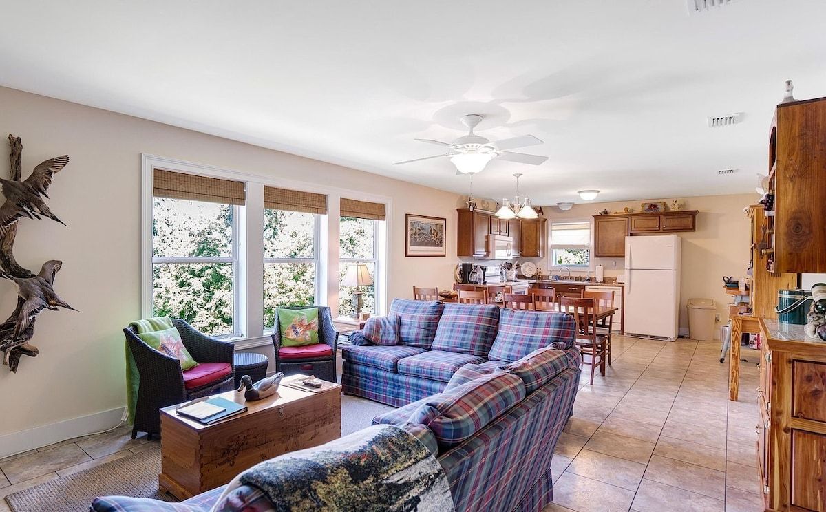 A spacious living area with a sofa, chairs, and a coffee table. There is a kitchen in the background with cabinets and a refrigerator.