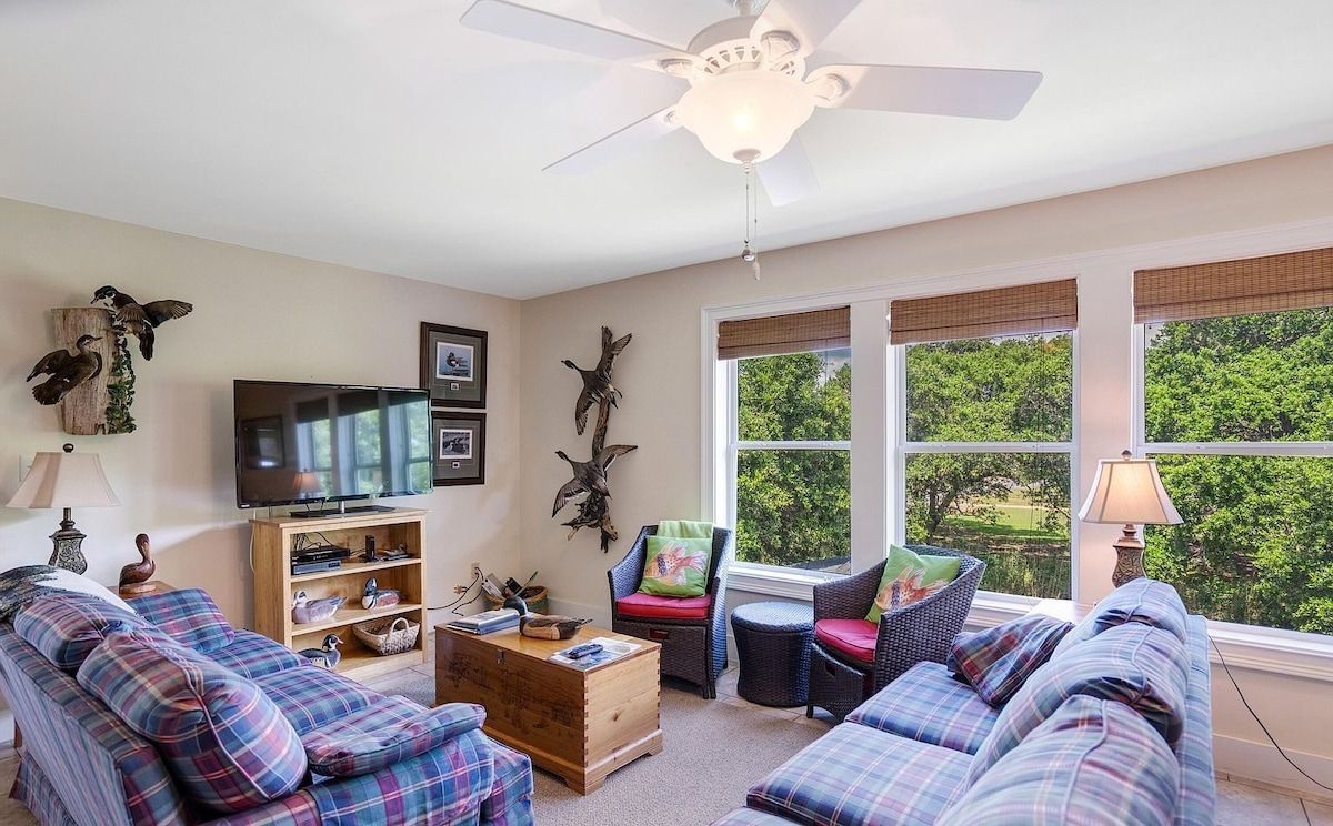 A living room with a ceiling fan, a television, and a large window with a view of greenery.