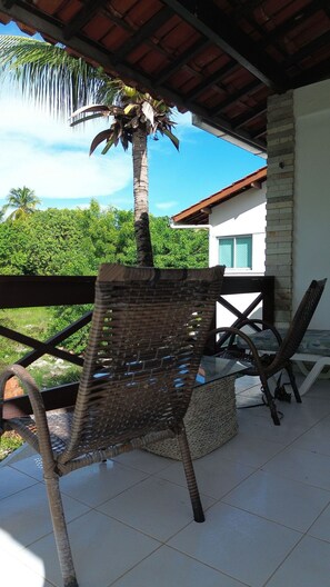 A covered outdoor seating area with wicker chairs, a palm tree, and a view of a house and greenery. - Privê das ondas Casa 6 (Ipojuca)