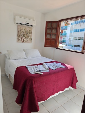 A bedroom with a bed, a red blanket, a white pillow, a wall-mounted air conditioner, and a window with a view of buildings. - Privê das ondas Casa 6 (Ipojuca)