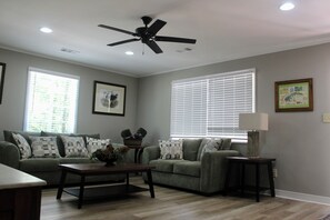 A living room with a ceiling fan, two windows with blinds, a sofa with patterned cushions, a coffee table, and a lamp on a side table. - The Dettor House  (Sardis)