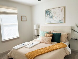 Una cama bien hecha con edredón blanco y beige, una manta amarilla y varios almohadones de distintos diseños y colores.