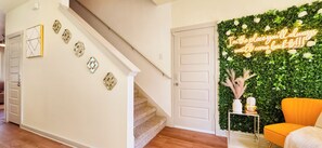 A hallway with a staircase, a door with a neon sign, and a wall decorated with greenery and white flowers. - Vrbo Property (San Marcos)