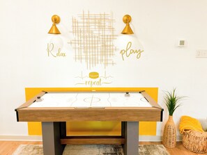 Una mesa de fútbol de mesa con diseño amarillo y blanco, una pared con un patrón geométrico y las palabras 'Relax' y 'play' en dorado, y una planta pequeña en una maceta de mimbre.