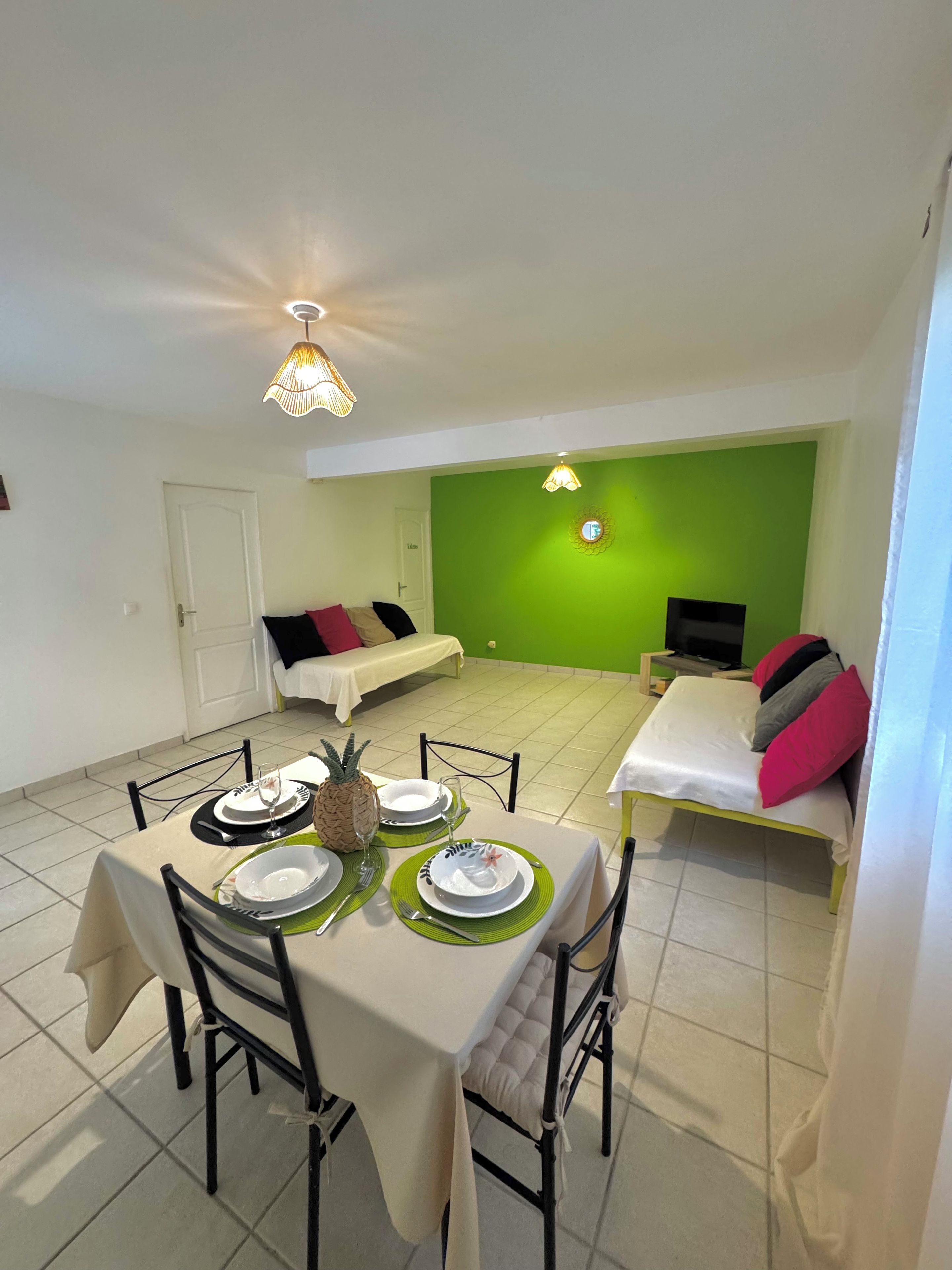 A dining area with a table set for two, a green wall, a bed with pillows, and a television in the background.