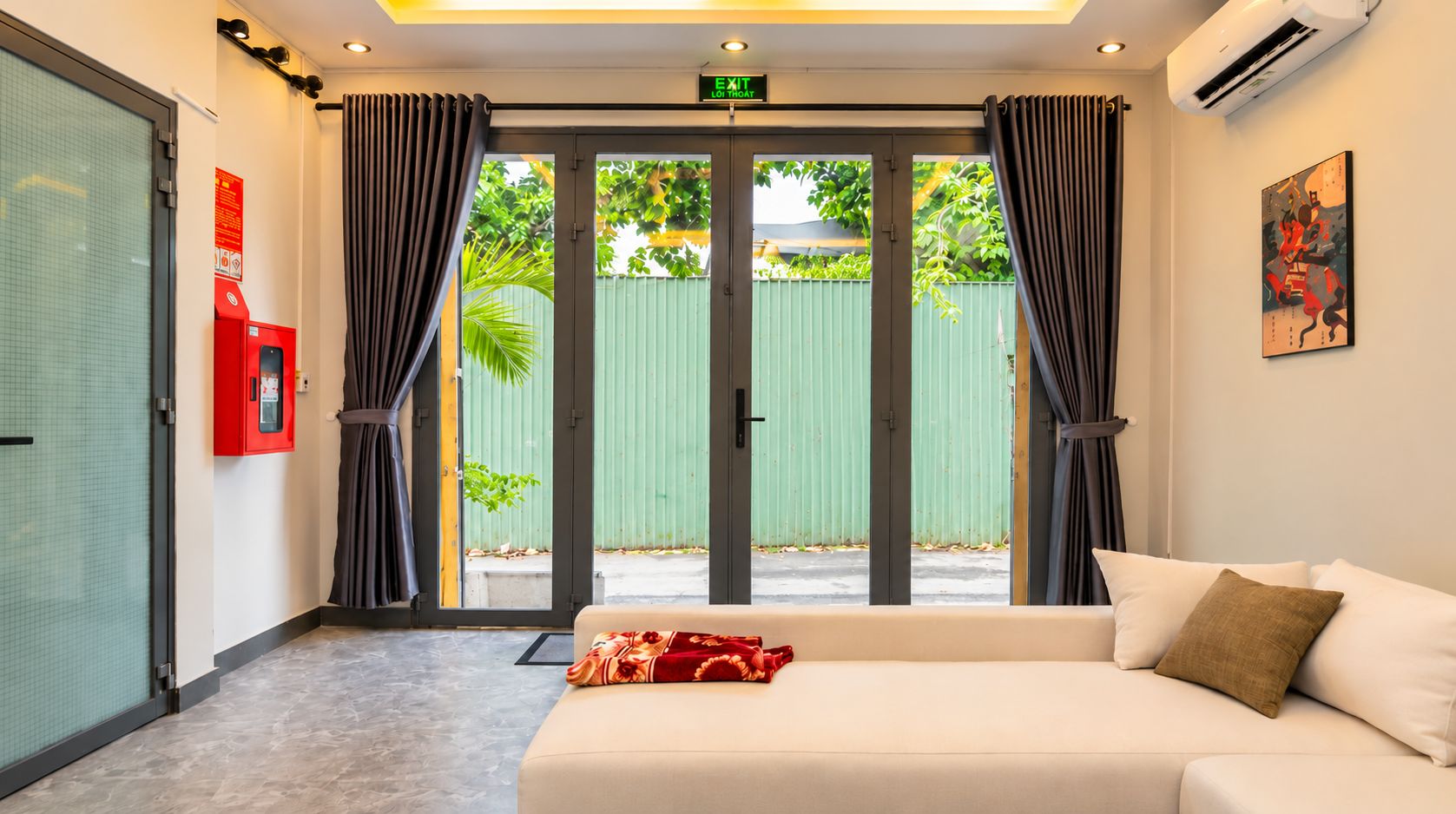 Ein Hotelzimmer mit Bett, Vorhängen, einer Klimaanlage, einem Feuerlöscher und Blick auf Grünflächen.