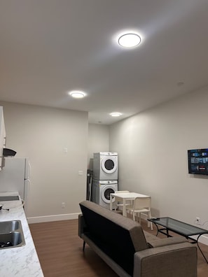 A compact living space with a sofa, a small table, a washing machine, and a TV mounted on the wall.