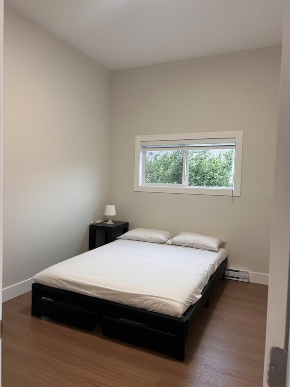 A bedroom with a single bed, a nightstand, a lamp, and a window with a view of greenery.