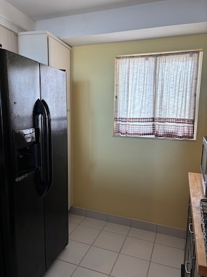 A kitchen with a refrigerator, a window with a checkered curtain, and tiled flooring. - Lovely 2-bedroom condo on the beach, fully furnished. (Satellite Beach)