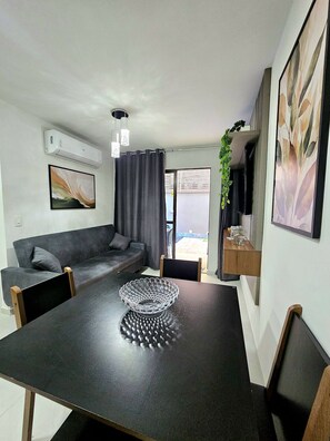 Ein modernes Wohnzimmer mit einem dunklen Sofa, einem Esstisch mit einer dekorativen Schale und einer Wandkonsole für eine Klimaanlage.