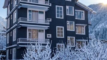 Un immeuble à plusieurs étages, doté de balcons et de fenêtres, entouré d’arbres couverts de neige et d’un mur de pierre.
