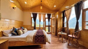 A wooden room with a bed, a table, and chairs, featuring large windows and a view of mountains.