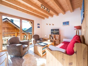 A cozy room with a wooden ceiling, a television, a sofa, and a view of mountains through large windows. - Chalet panoramic view - 9 guests (La Toussuire)