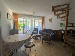A living room with a dining table, chairs, a sofa, a staircase, and a view through a window. - Seafront apartment, air conditioning - 7 guests (La Grande-Motte)