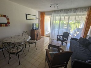 A living room with a dining table, chairs, a sofa, a television, and a window with curtains. - Seafront apartment, air conditioning - 7 guests (La Grande-Motte)