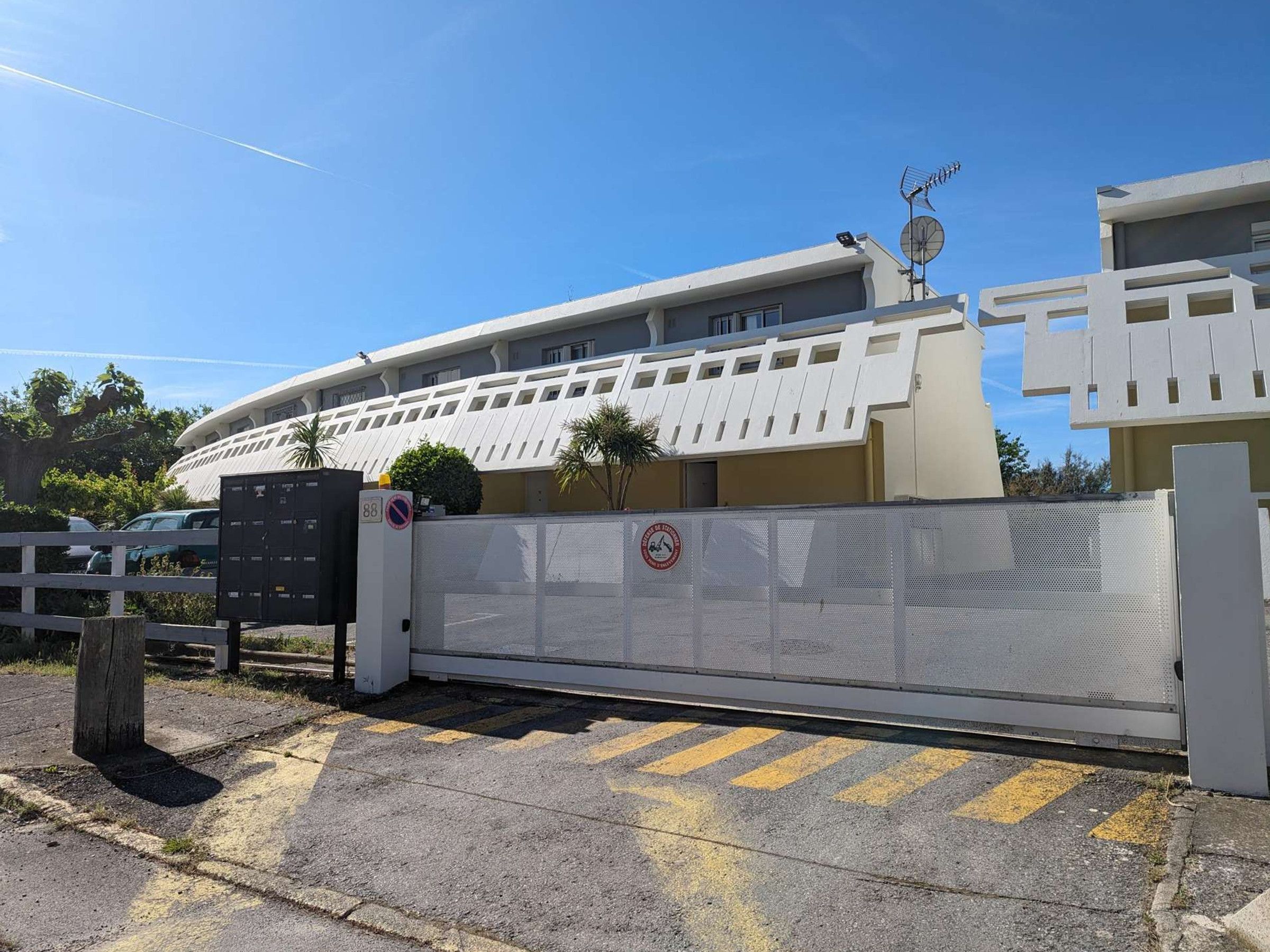 Une entrée fermée par un portail, agrémentée d’un panneau interdisant le stationnement, un bâtiment à l’architecture moderne et un ciel dégagé.