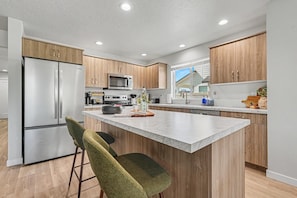 A modern kitchen with wooden cabinets, a stainless steel refrigerator, a microwave, and a large island with bar stools. - 5BR/3BA Gem State Getaway | Game Room + Yard (Nampa)