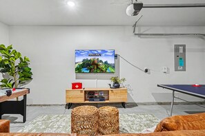 A room with a television mounted on the wall, a ping pong table, a foosball table, and a wooden cabinet with a red first aid kit. - 5BR/3BA Gem State Getaway | Game Room + Yard (Nampa)