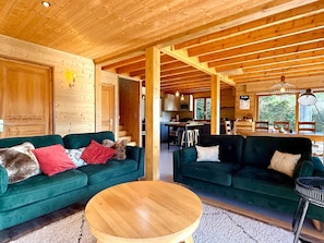 A cozy cabin interior with two green sofas, a wooden dining table, and a kitchen area visible through an open doorway. - Renovated chalet, exceptional views - 8 guests (Les Gets)