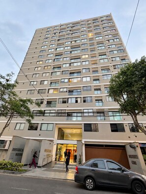 Um edifício de vários andares com design arquitetônico moderno, apresentando entrada no nível da rua com portas de vidro e área de estacionamento visível.